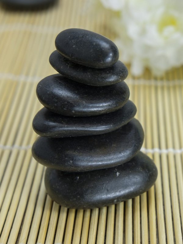 Aufgestapelte schwarze Massagesteine neben einer weißen Blüte auf einem Bambusuntergrund.