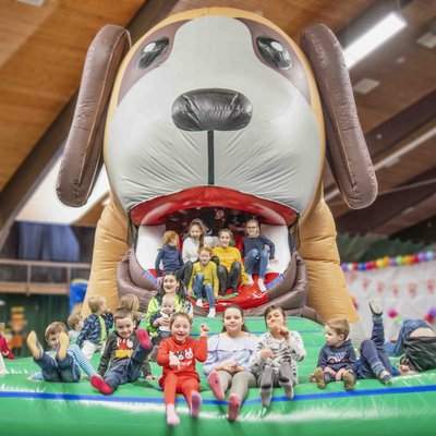 Eine Gruppe fröhlicher Kinder spielt auf einer riesigen Hüpfburg in Form eines Hundekopfes in der Indoor-Spielewelt des Hotels Vier Jahreszeiten.