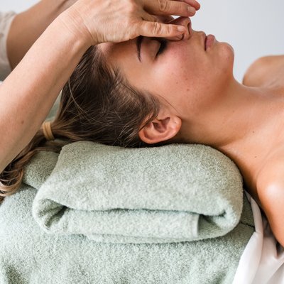 Frau liegt auf einer Massageliege auf dem Rücken und wird von zwei Händen im Gesicht massiert