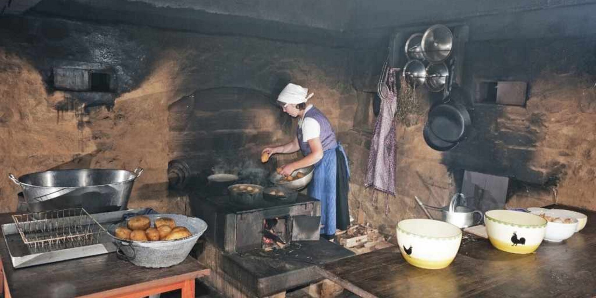 Blick in eine rustikale, historische Küche mit offenem Kamin und alten Kochutensilien.