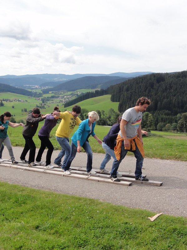Mehrere Personen stehen auf einem sehr langen Ski und fahren damit über einen Schotterweg - Schwarzwaldgaudi