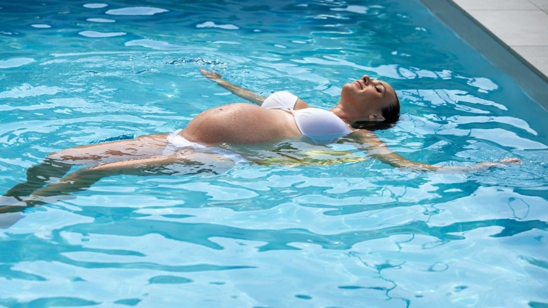 Eine schwangere Frau lässt sich entspannt auf dem Rücken im klaren blauen Wasser eines Pools treiben.