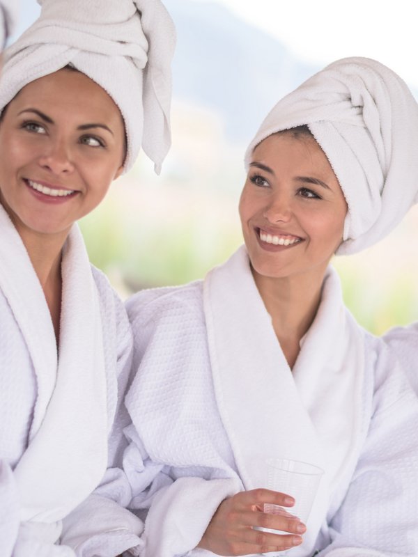 Vier lächelnde Frauen in weißen Bademänteln unterhalten sich angeregt in einem hellen Wellnessbereich.