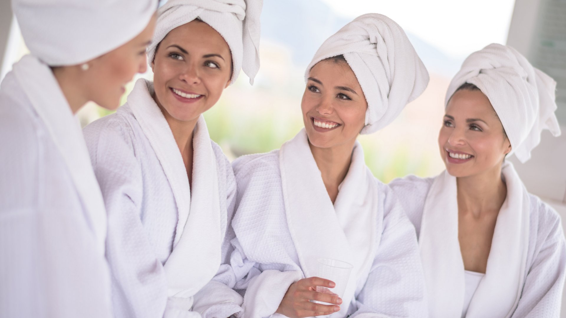 Vier lächelnde Frauen in weißen Bademänteln unterhalten sich angeregt in einem hellen Wellnessbereich.