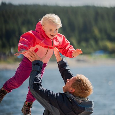 Familienmoment im Urlaub: Ein Vater hebt sein Kind freudig in die Luft vor der Seekulisse.