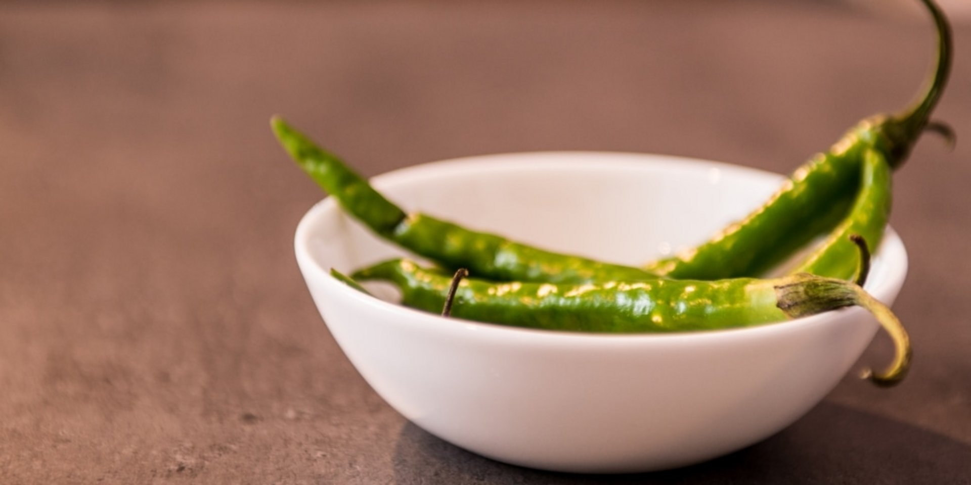 Eine kleine weiße Schale mit frischen grünen Chilischoten auf einer Schieferplatte.