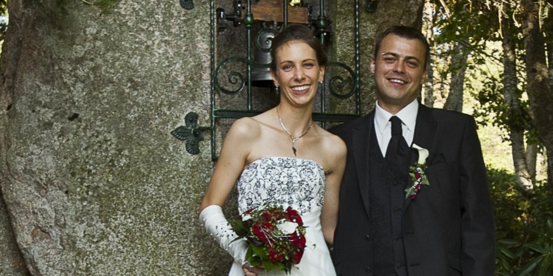 Ein Brautpaar posiert für ein festliches Foto vor einer Steinmauer mit Gästen im Vordergrund.