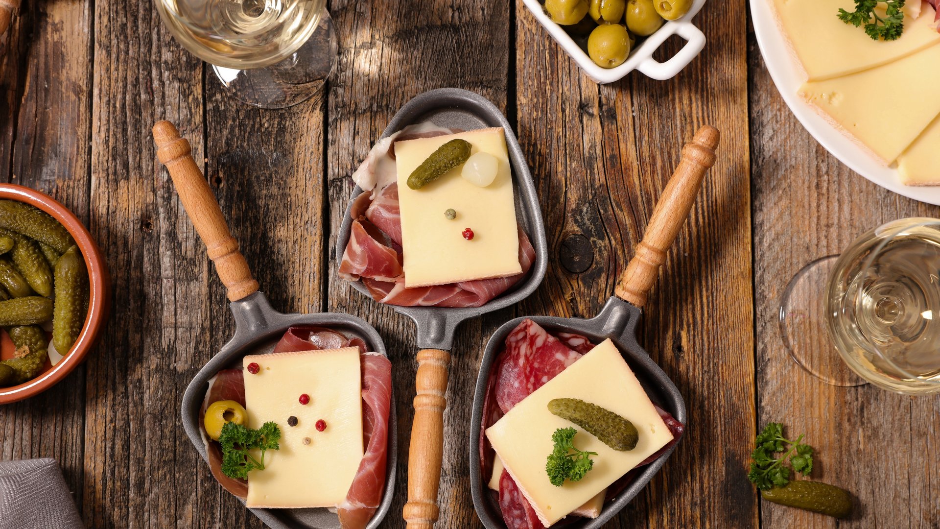 Auf einem Tisch stehen drei Raclettepfannen mit Käse, Tomate, Schinken und Kräutern, Wein, Oliven und Gurken