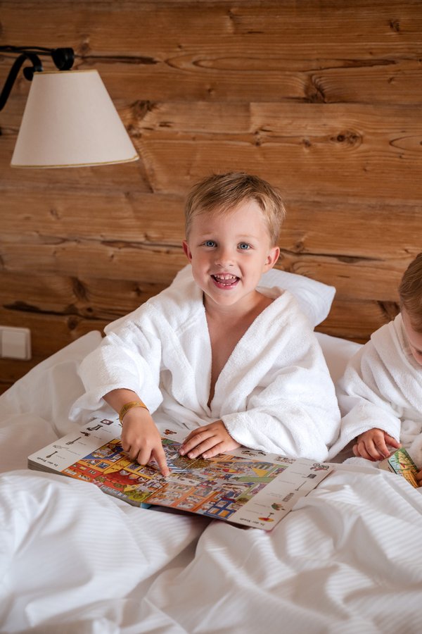 Ein Kind im weißen Bademantel sitzt mit einem Buch im gemütlichen Hotelbett des Hotels Vier Jahreszeiten im Schwarzwald.