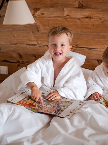 Ein Kind im weißen Bademantel sitzt mit einem Buch im gemütlichen Hotelbett des Hotels Vier Jahreszeiten im Schwarzwald.