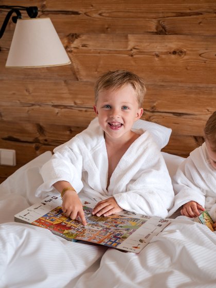 Ein Kind im weißen Bademantel sitzt mit einem Buch im gemütlichen Hotelbett des Hotels Vier Jahreszeiten im Schwarzwald.