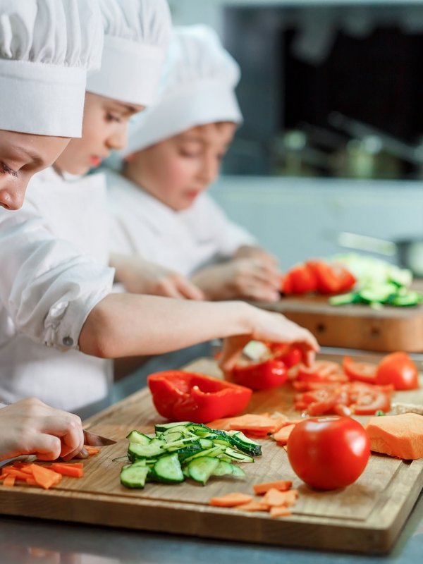 Eine Gruppe von Kindern in Kochmützen bereitet mit frischen Zutaten ein Menü im Hotel Vier Jahreszeiten zu.
