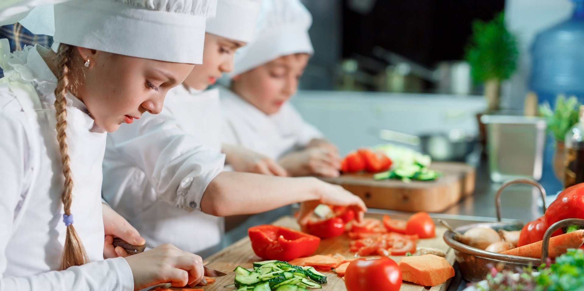 Eine Gruppe von Kindern in Kochmützen bereitet mit frischen Zutaten ein Menü im Hotel Vier Jahreszeiten zu.