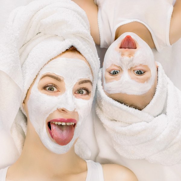 Zwei Frauen in weißen Bademänteln und Handtuch-Turbans lachen mit aufgetragenen Gesichtsmasken.