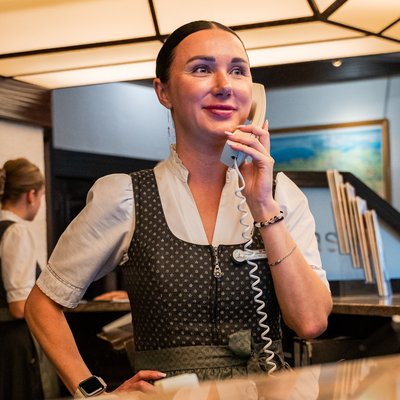 Eine lächelnde Mitarbeiterin am Telefon im Servicebereich des Hotels Vier Jahreszeiten kümmert sich um die Wünsche der Gäste.