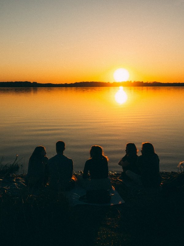 Eine Gruppe von Menschen sitzt bei Sonnenuntergang am Seeufer und genießt die gemeinsame Zeit.