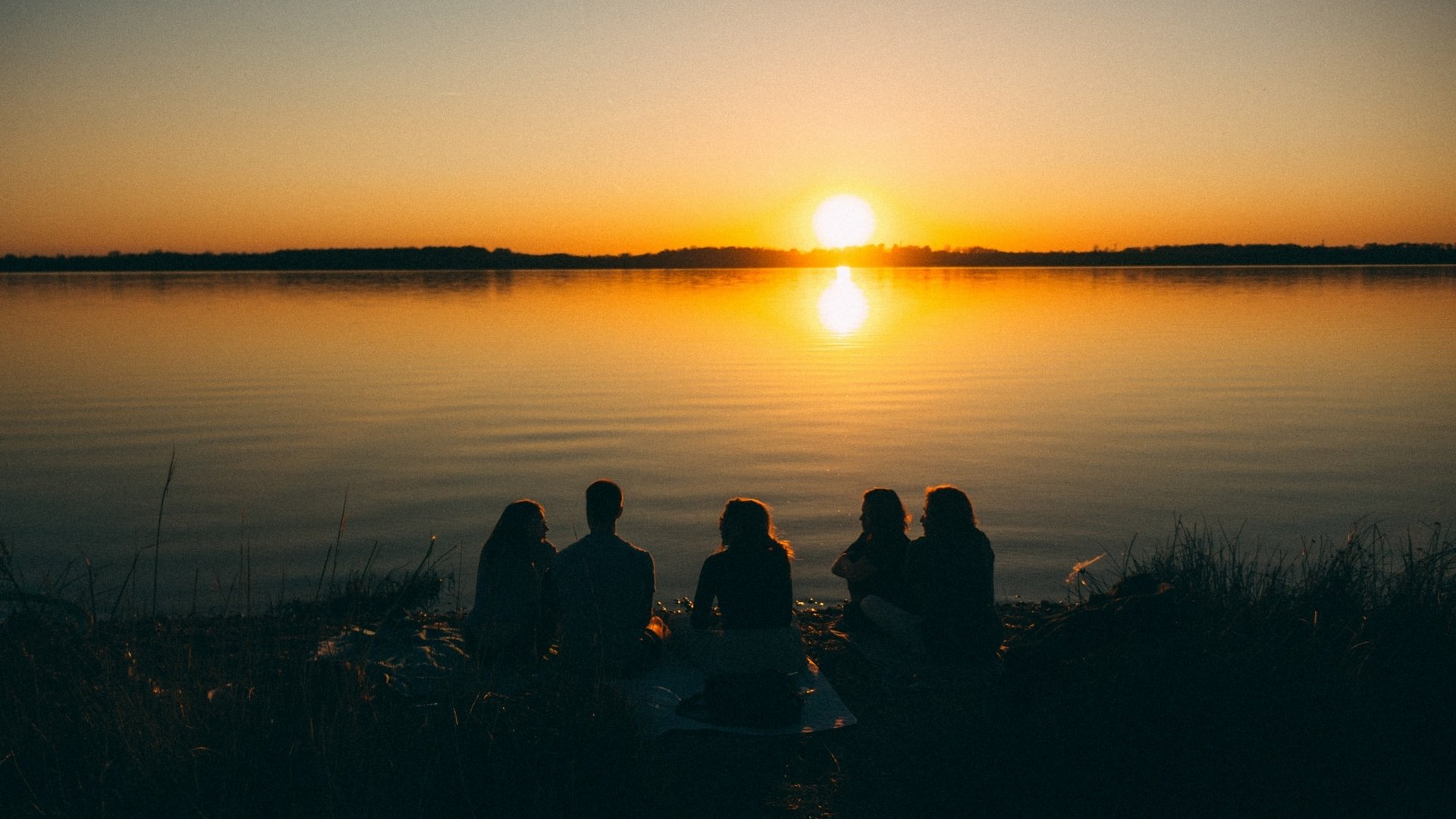 Eine Gruppe von Menschen sitzt bei Sonnenuntergang am Seeufer und genießt die gemeinsame Zeit.