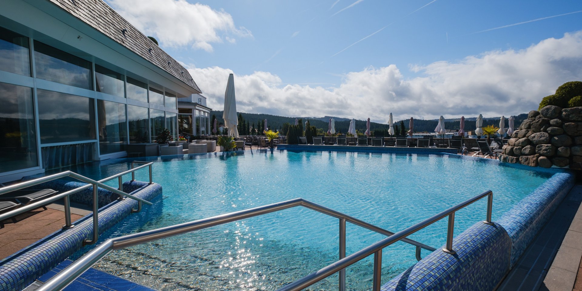 Beheizter Außenpool mit Blick auf die umliegende Natur und Liegeflächen unter blauem Himmel.