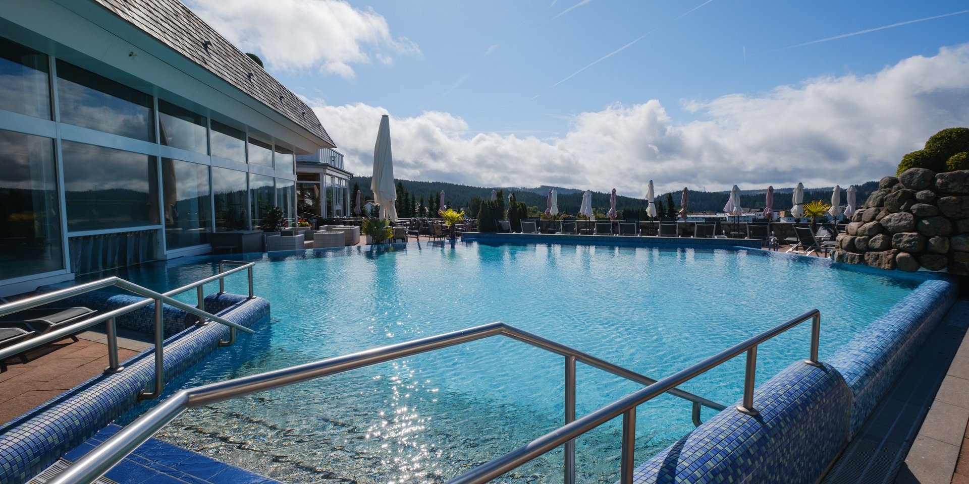 Beheizter Außenpool mit Blick auf die umliegende Natur und Liegeflächen unter blauem Himmel.