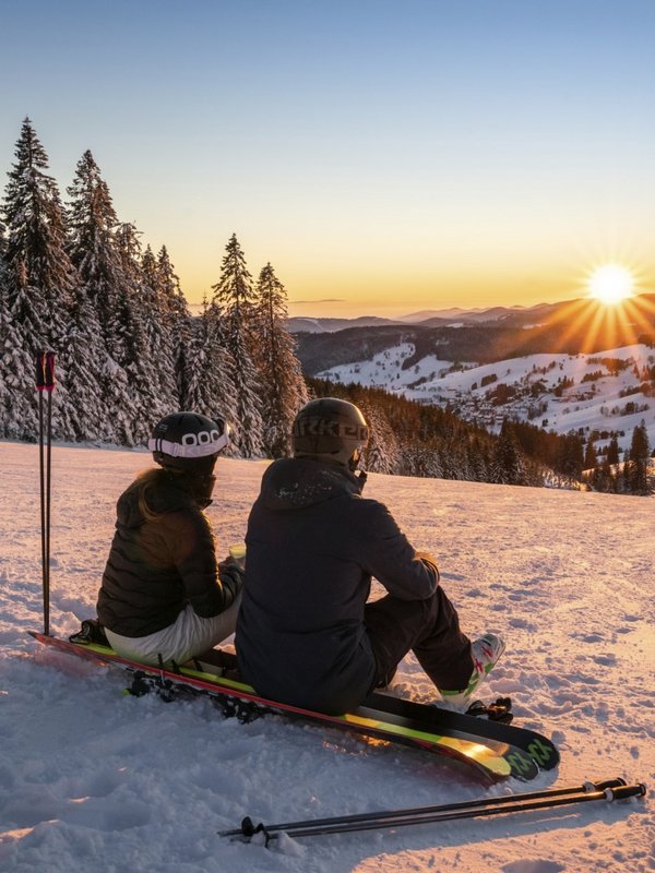 Zwei Personen genießen bei einer Pause auf ihren Snowboards den Sonnenuntergang im Skigebiet.