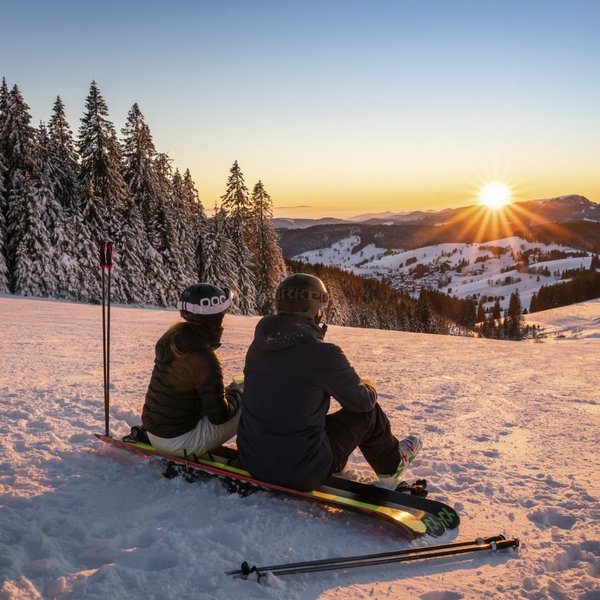 Zwei Personen genießen bei einer Pause auf ihren Snowboards den Sonnenuntergang im Skigebiet.