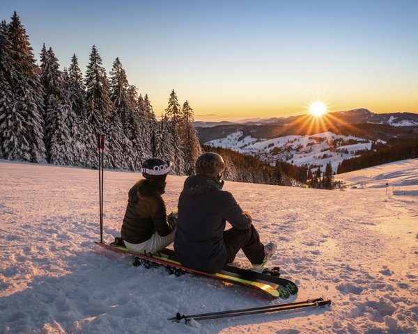Zwei Personen genießen bei einer Pause auf ihren Snowboards den Sonnenuntergang im Skigebiet.