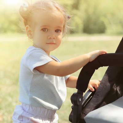 Ein kleines Mädchen schiebt einen Spielzeug-Kinderwagen über eine weite, grüne Wiese.