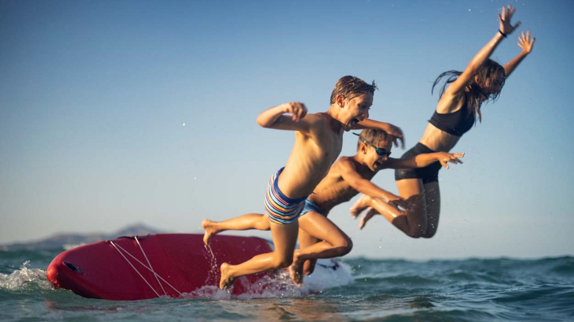 Drei Kinder springen gleichzeitig mit viel Freude von einem SUP.