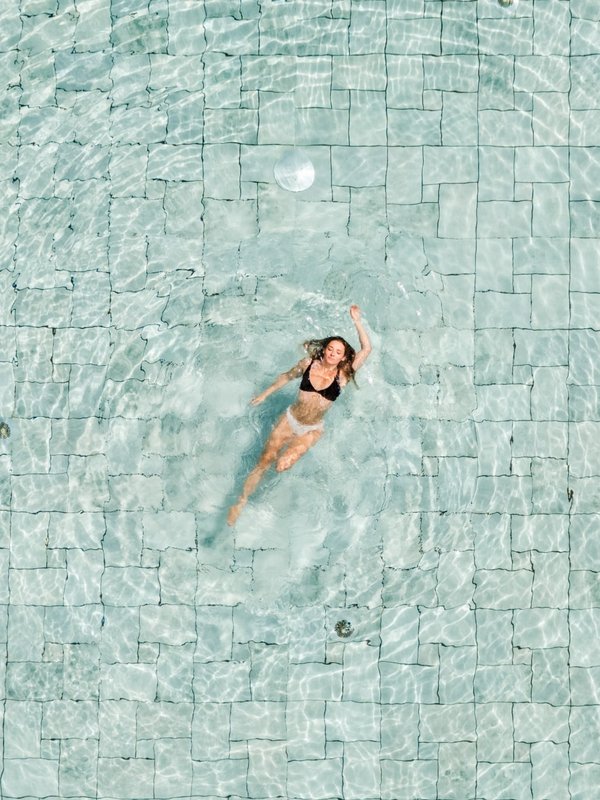 Luftaufnahme einer Person, die entspannt im glasklaren Wasser des hoteleigenen Außenpools im Schwarzwald treibt.