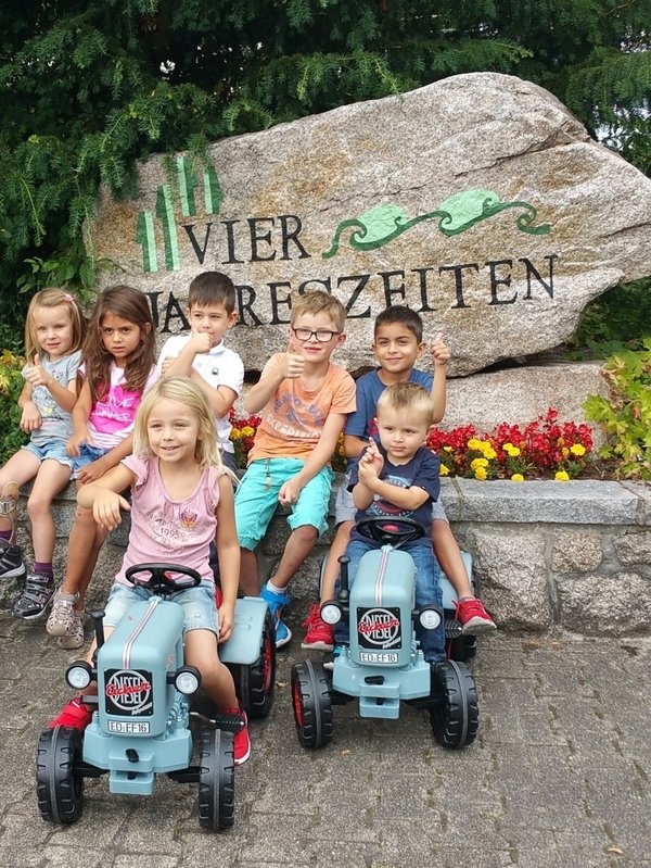 Eine Gruppe fröhlicher Kinder spielt auf blauen Trettraktoren vor dem großen Namensstein des Hotels Vier Jahreszeiten im Schwarzwald.