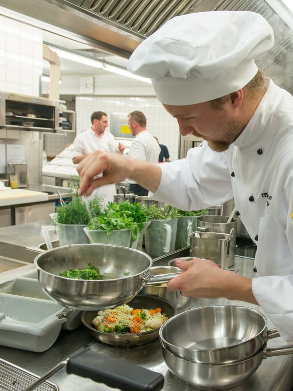 Ein professioneller Koch in weißer Uniform richtet konzentriert Speisen in der Küche des Hotels Vier Jahreszeiten an.