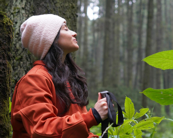 Frau steht im Wald mit geschlossenen Augen, sie trägt eine Mütze und eine rote Jacke