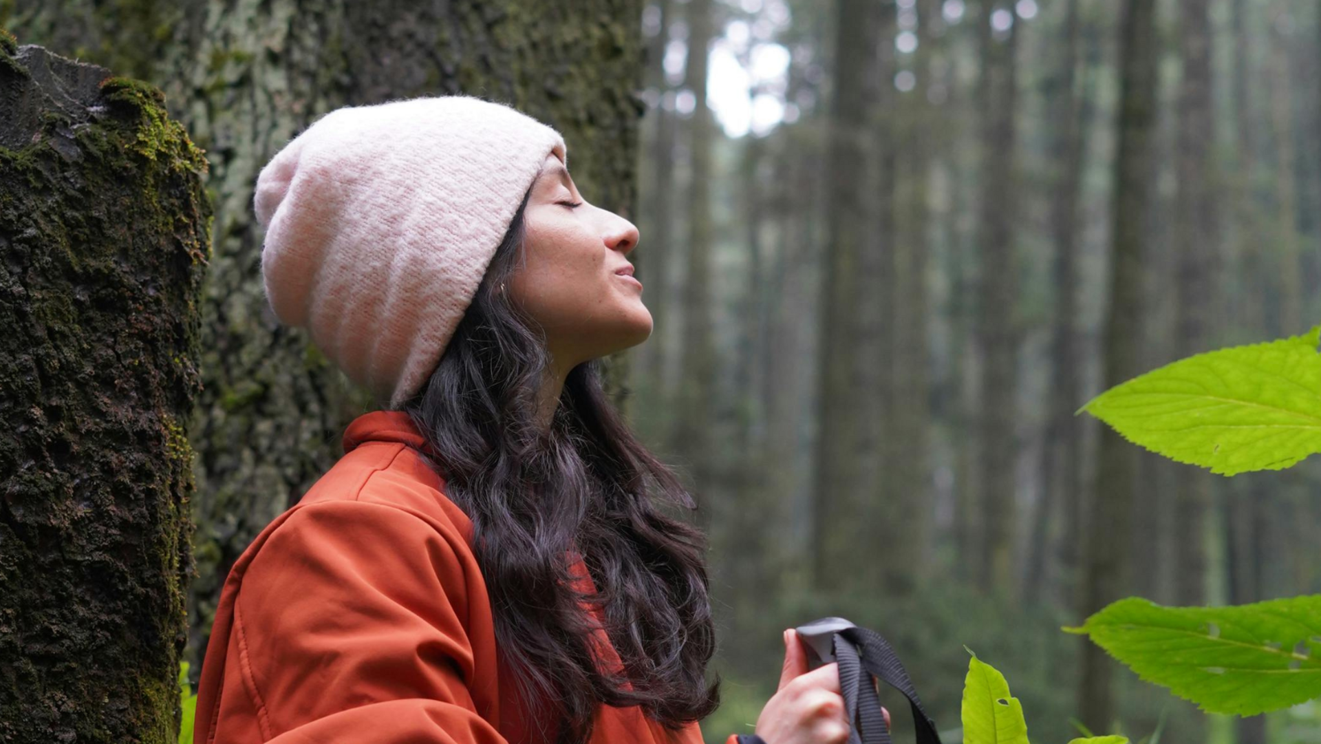 Frau steht im Wald mit geschlossenen Augen, sie trägt eine Mütze und eine rote Jacke