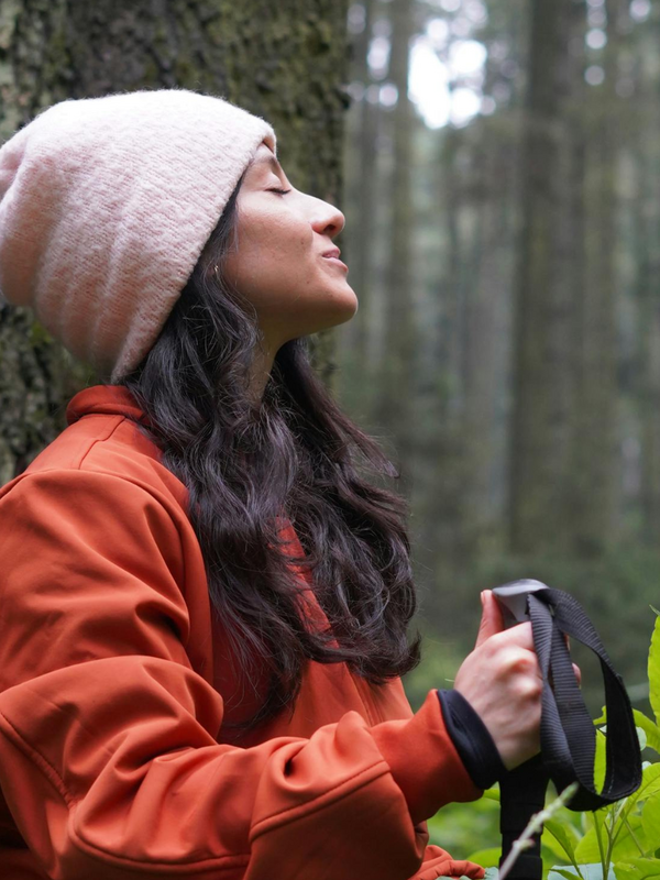 Frau steht im Wald mit geschlossenen Augen, sie trägt eine Mütze und eine rote Jacke