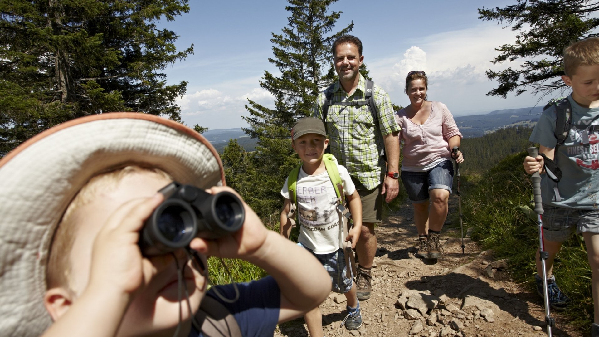 Eine Familie wandert mit Rucksäcken über eine sonnige Anhöhe mit weitem Ausblick.