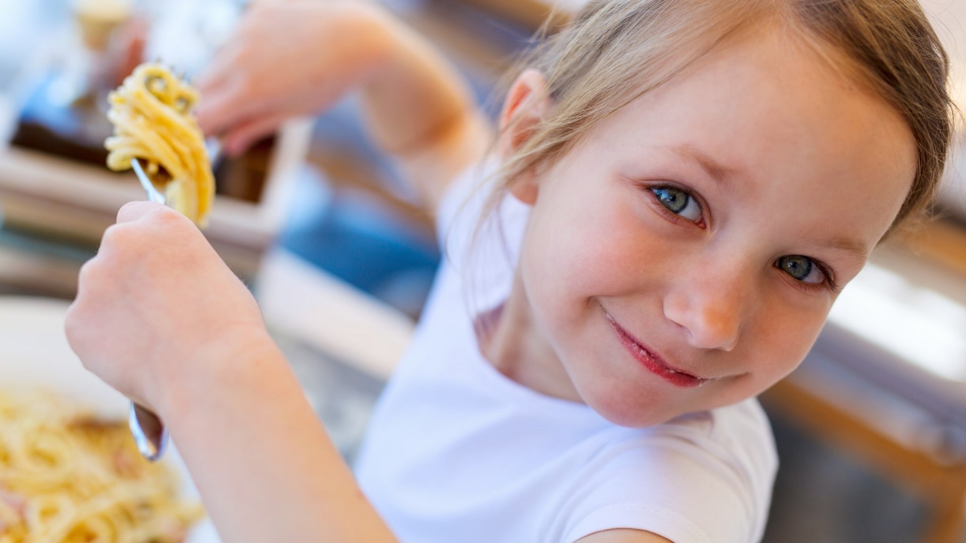 Porträt eines lächelnden kleinen Mädchens, das eine Gabel mit Nudeln hochhält.