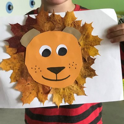 Ein Kind hält ein selbstgebasteltes Bild aus bunten Herbstblättern, das ein Löwengesicht zeigt.