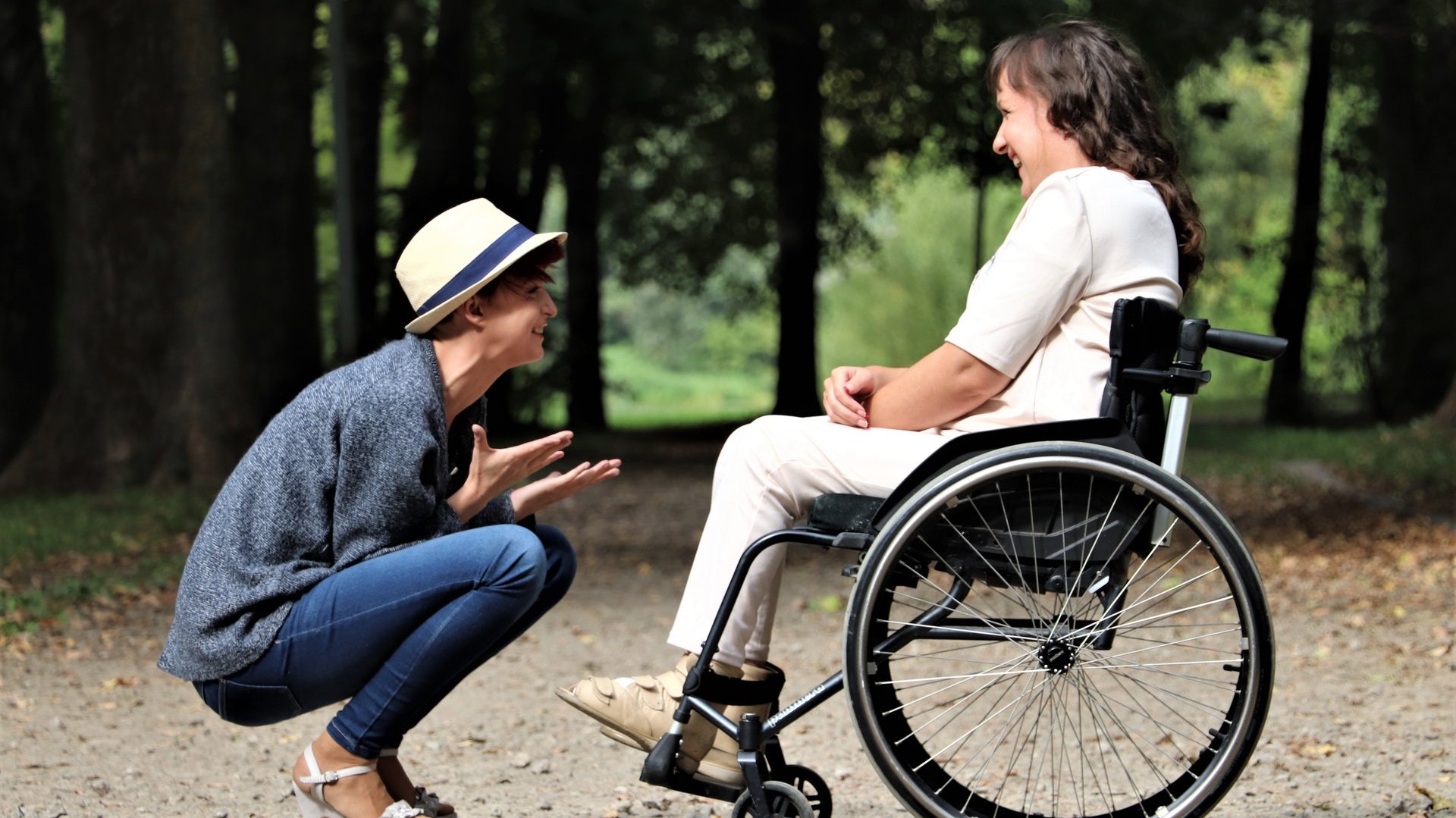 Eine Frau kniet im Wald vor einer Frau im Rollstuhl und hält ihre Hände in einer herzlichen Geste.