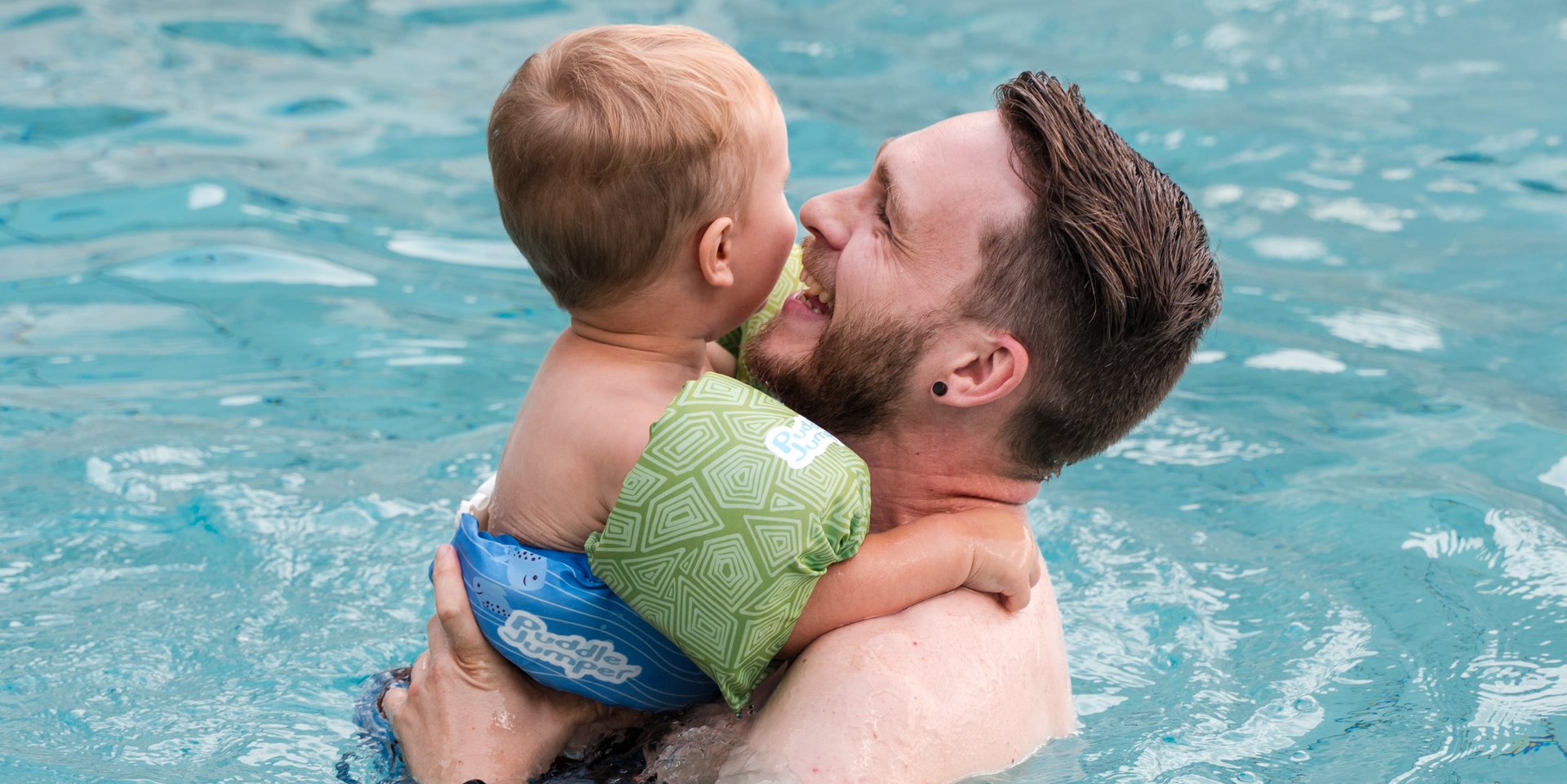 Ein Vater hebt sein lachendes Kleinkind im flachen Wasser des VJZ Hotelpools hoch.