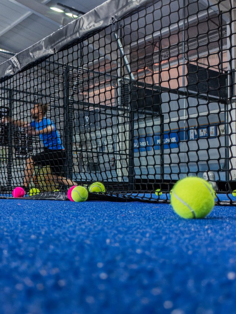 Blick über das Netz auf einen blauen Indoor-Tennisplatz mit einem Spieler im Hintergrund.