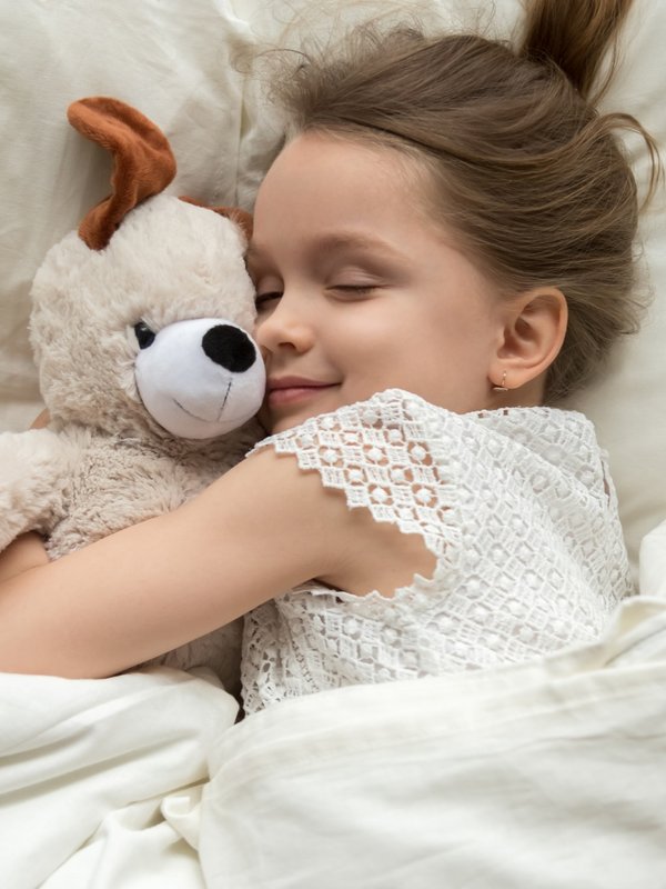 Ein kleines Mädchen schläft friedlich in weißen Kissen und kuschelt mit einem Teddybären.