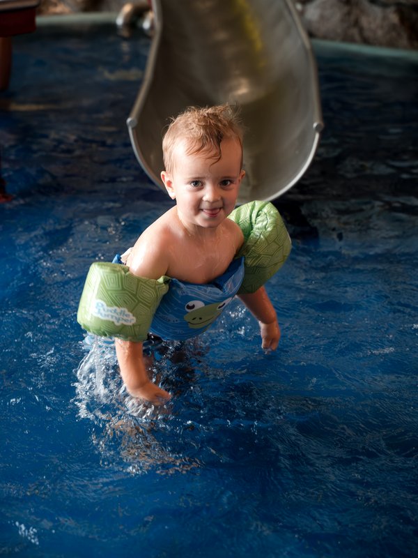 Kleinkind steht vor einer Rutsche im Wasser und trägt Schwimmflügel