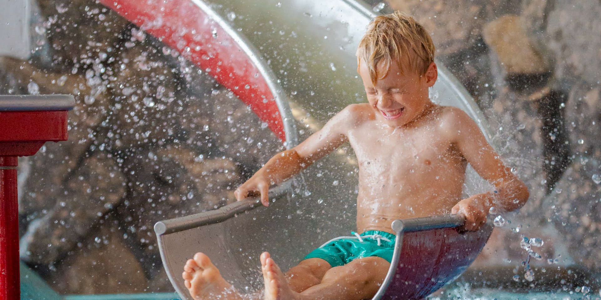 Ein kleiner Junge rutscht mit Schwung über eine Kinderrutsche direkt ins Wasserbecken des Hotels Vier Jahreszeiten.