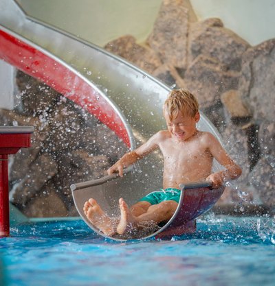 Ein kleiner Junge rutscht mit Schwung über eine Kinderrutsche direkt ins Wasserbecken des Hotels Vier Jahreszeiten.