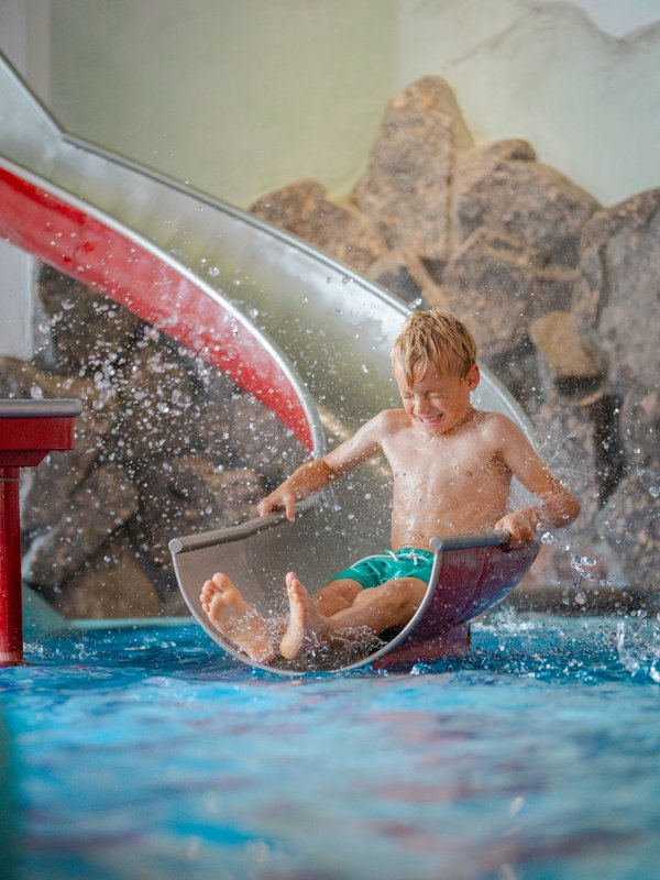 Ein kleiner Junge rutscht mit Schwung über eine Kinderrutsche direkt ins Wasserbecken des Hotels Vier Jahreszeiten.