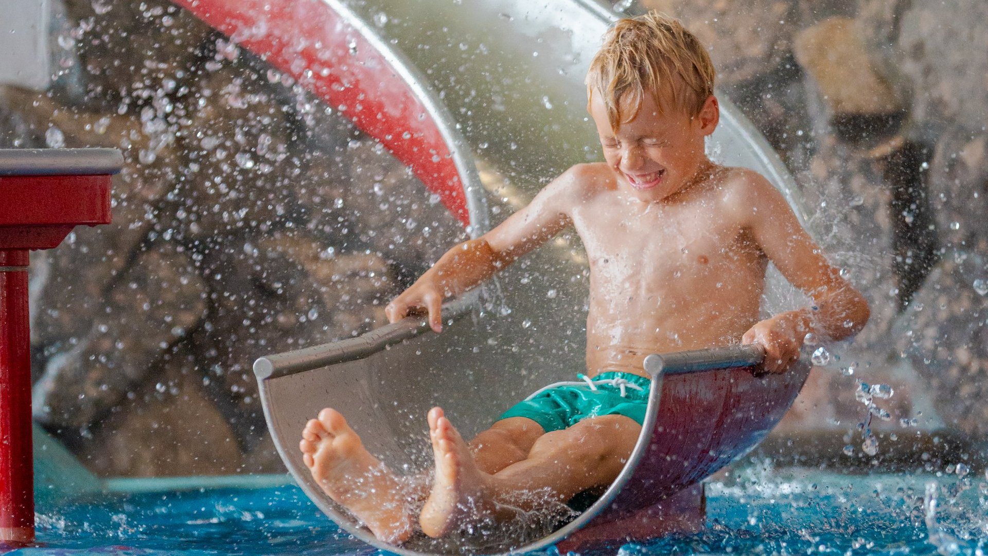 Ein kleiner Junge rutscht mit Schwung über eine Kinderrutsche direkt ins Wasserbecken des Hotels Vier Jahreszeiten.