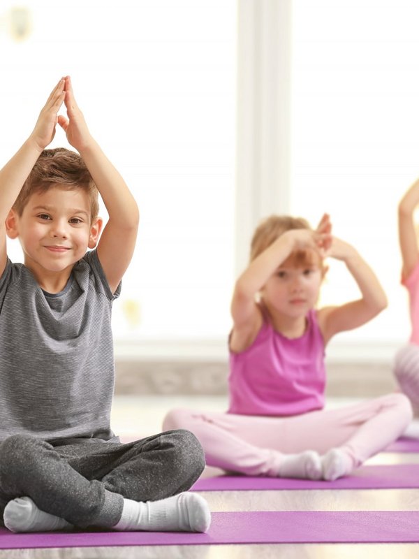 Drei Kinder machen gemeinsam spielerische Yoga-Übungen in einem hellen Gymnastikraum.
