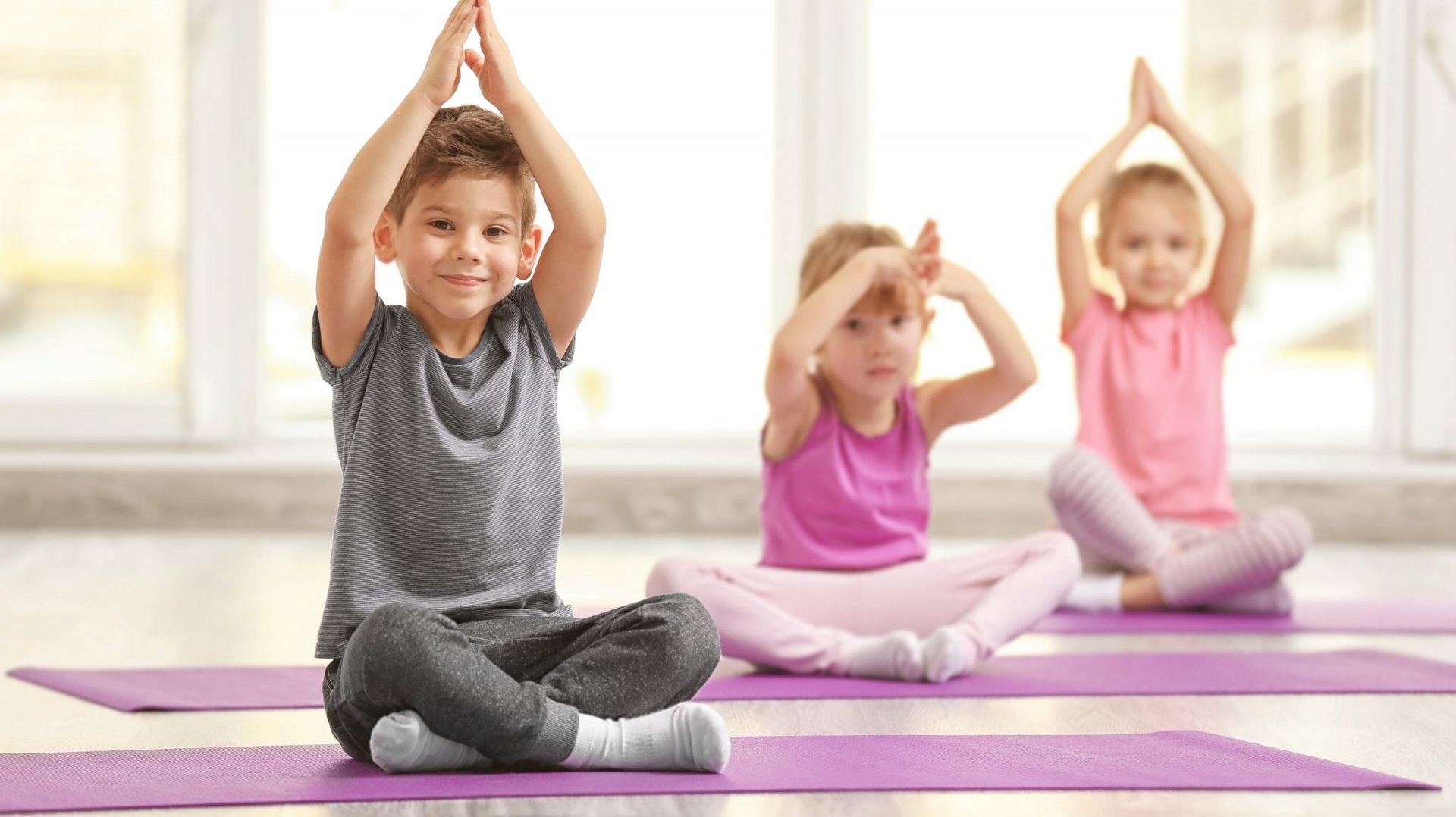 Drei Kinder machen gemeinsam spielerische Yoga-Übungen in einem hellen Gymnastikraum.