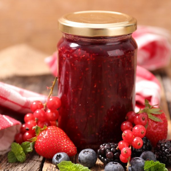Ein Glas hausgemachte Beerenmarmelade neben frischen Früchten auf einem Holztisch.