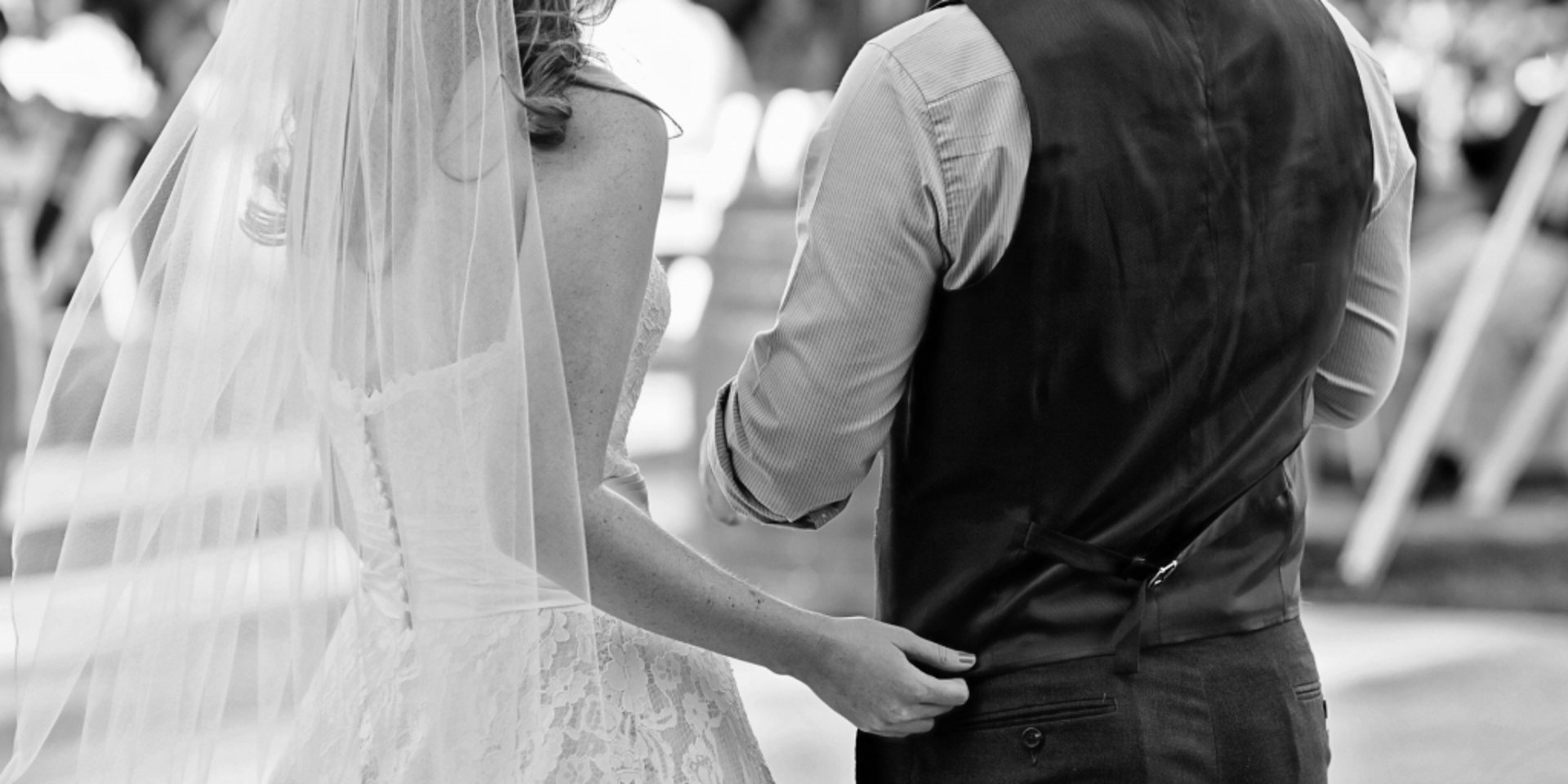 Rückenansicht in Schwarz-Weiß eines Brautpaares beim gemeinsamen Spaziergang durch einen festlich dekorierten Garten.